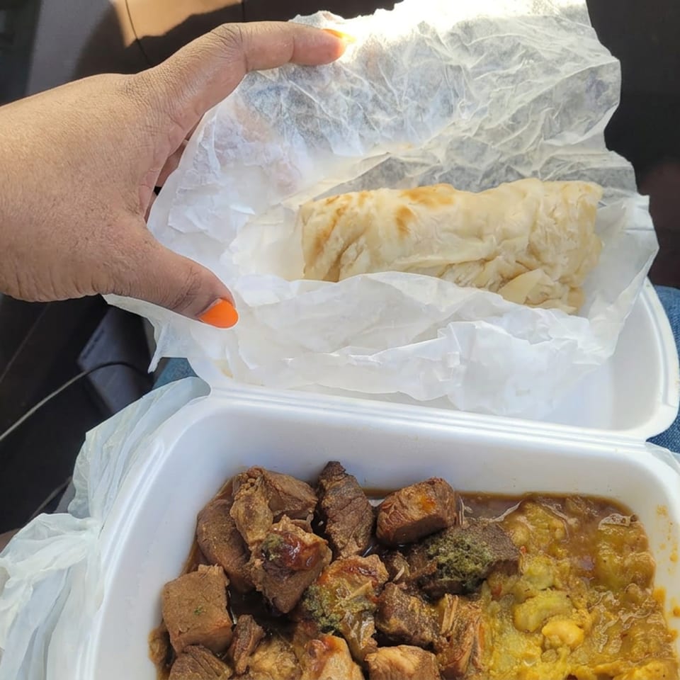 Open takeout container holding browned meat and yellow stew, with a flaky pastry in wax paper, inside a car.